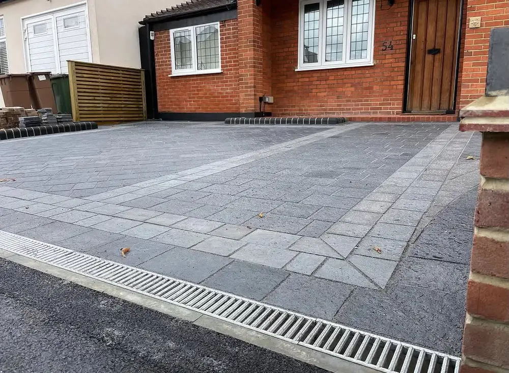 Grey patterned driveway paving with light border bands, raised entrance edging and a drainage channel along the front